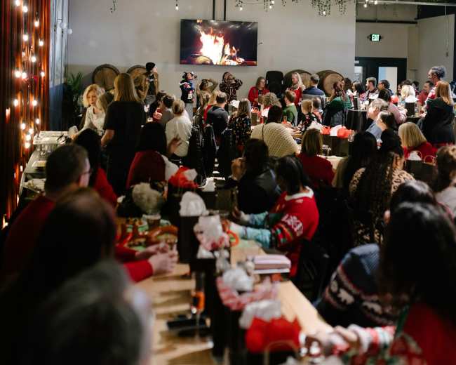 A crowded restaurant features guests enjoying a festive gathering with a visible fireplace on a screen and holiday decorations overhead.