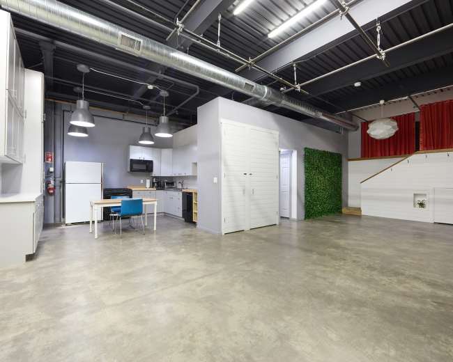 The image shows a spacious, modern interior with a kitchen area, a dining table, and a partially decorated wall featuring greenery.