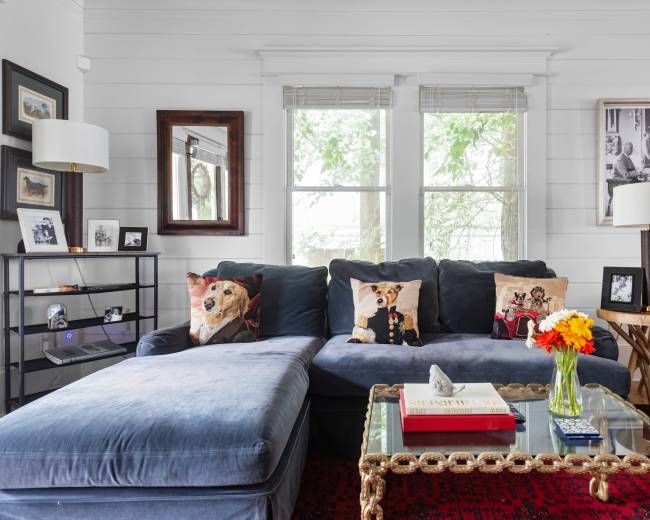 The living room features a blue sectional sofa adorned with decorative pillows, a glass coffee table, and various framed artworks on the white walls.