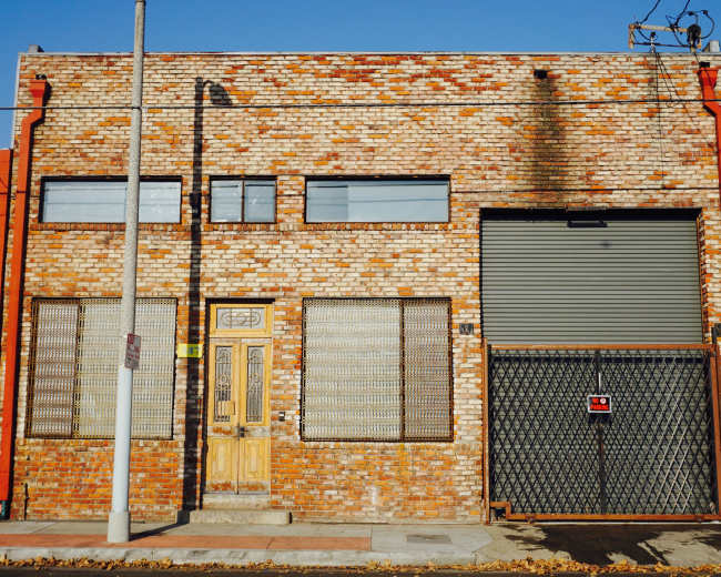 The image shows a brick industrial building with large windows, a wooden door, and a metal security gate.