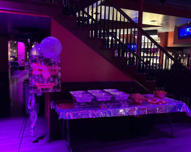 A decorated table with food trays and balloons is positioned near a staircase in a dimly lit venue.