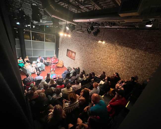 A diverse audience watches a performance on stage in a dimly lit venue with exposed brick walls.