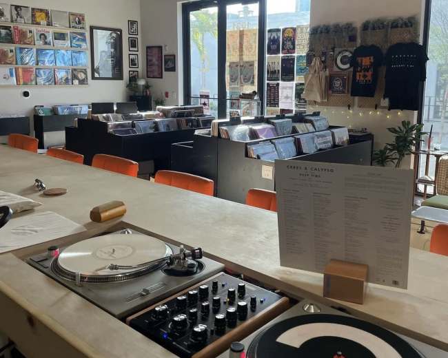 The image shows a record store with a bar area featuring turntables, a menu, and shelves filled with vinyl records in the background.