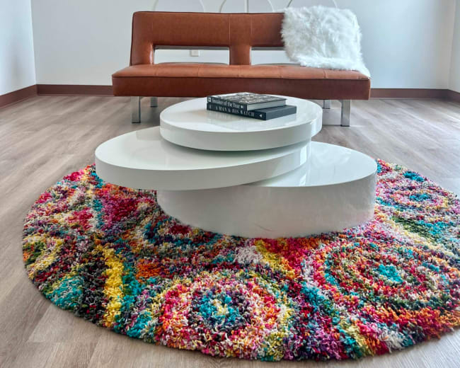 The image shows a modern living space featuring a brown sofa, a circular multicolored rug, and a white coffee table on hardwood flooring.