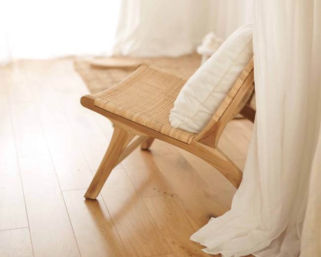 A wooden chair with a white cushion is positioned beside sheer curtains and a light-colored rug in a spacious, well-lit room.