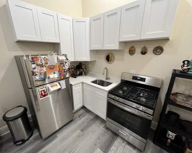 The image shows a small kitchen with white cabinets, a stainless steel refrigerator, a stainless steel stove, and a sink in the corner.