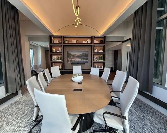 A large, oval conference table surrounded by white chairs is positioned in a brightly lit meeting room with a scenic backdrop and bookshelves.