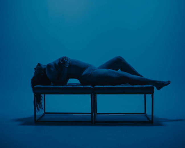 A person lies on a bench against a blue background, with their body arched and arms crossed over their chest.