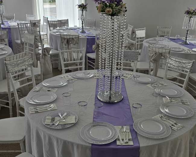 The image shows a banquet hall set for a formal event, featuring round tables covered with white tablecloths and lavender runners, adorned with crystal centerpieces and floral arrangements.