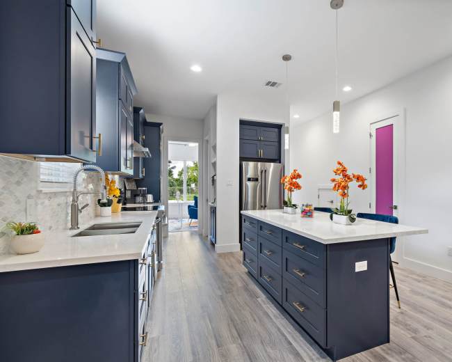 The image shows a modern kitchen with dark blue cabinetry, a central island with a white countertop, and a light-filled space featuring vibrant purple doors.