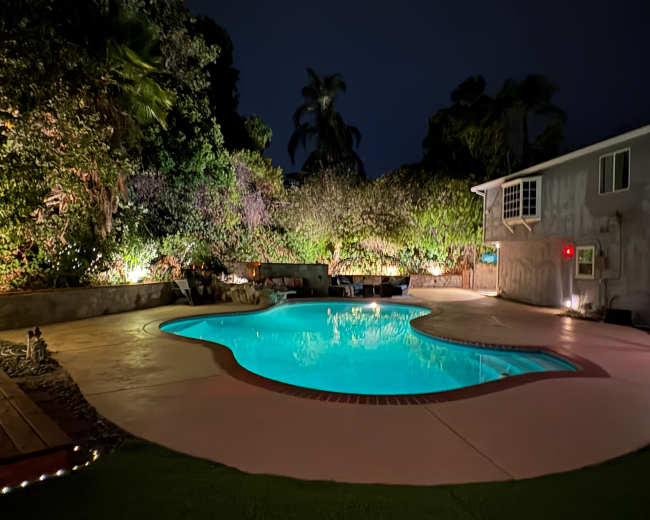 The image shows a backyard pool area at night, illuminated by soft lights, surrounded by lush greenery and a house to one side.