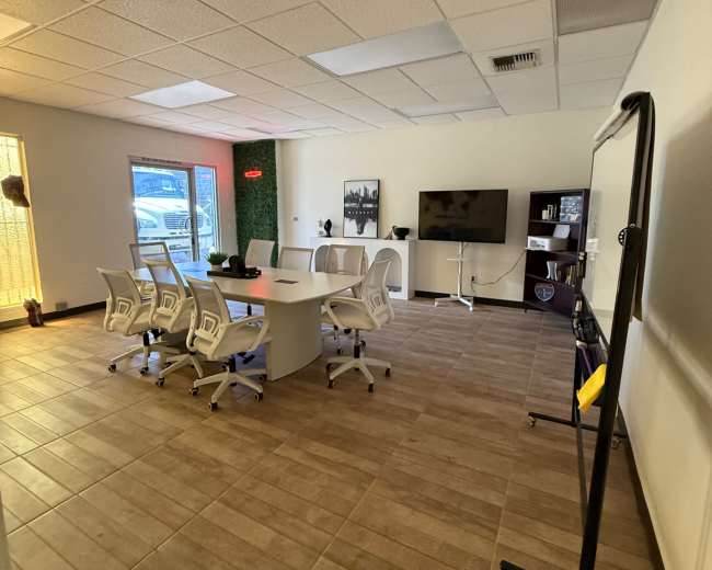 The image shows a conference room with a large round table surrounded by several white office chairs, a television mounted on the wall, and a green accent wall.