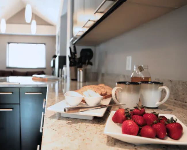 A kitchen countertop displays a plate of strawberries, a small dish with cream, cookies, and a bottle of honey, with a blurred living area visible in the background.