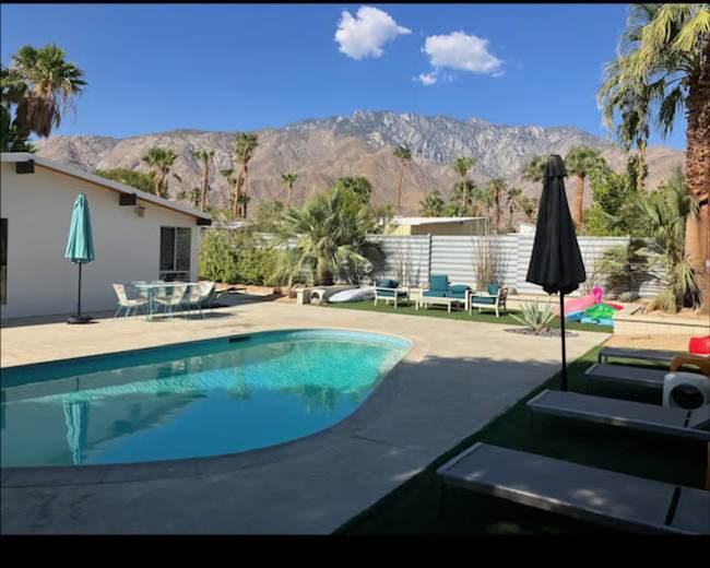 A swimming pool is located in a backyard surrounded by palm trees and mountains, with patio furniture visible nearby.