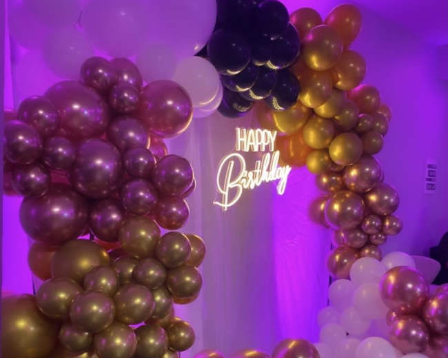 A decorative backdrop for a birthday celebration featuring an arrangement of balloons in gold, black, and white, with a illuminated "Happy Birthday" sign.