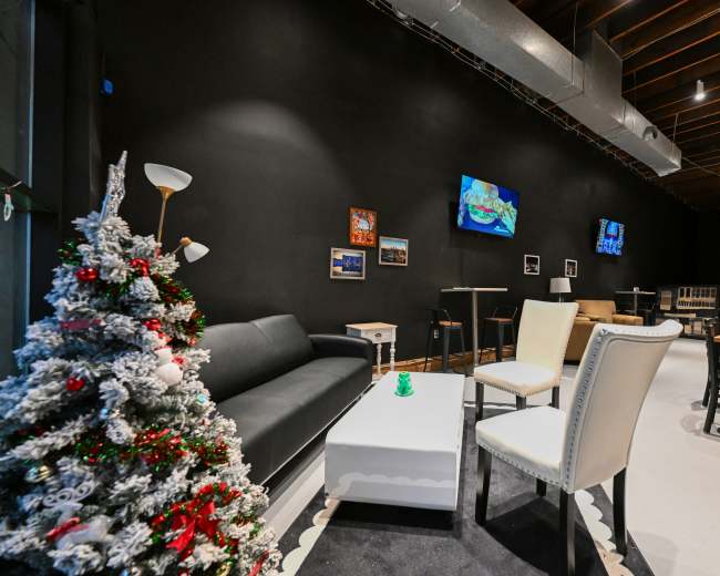 The scene features a modern lounge area with black walls, a decorative Christmas tree, a white coffee table, seating, and flat-screen TVs mounted on the walls.
