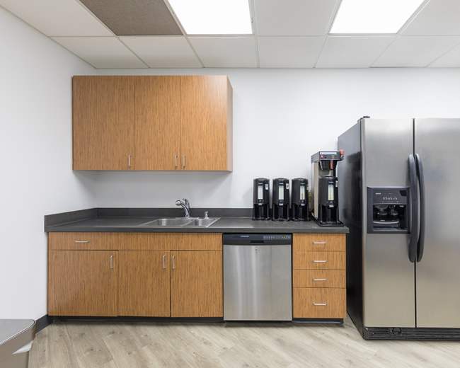 The image shows a modern kitchen area featuring a stainless steel refrigerator, a dishwasher, a sink, and several coffee machines against a wooden cabinet backdrop.