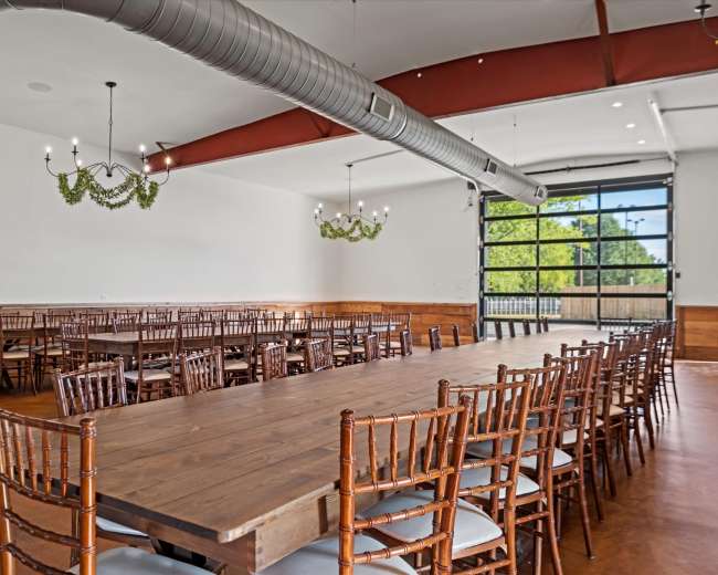 The image shows a spacious dining area with multiple long wooden tables and wooden chairs, featuring large windows and decorative light fixtures.