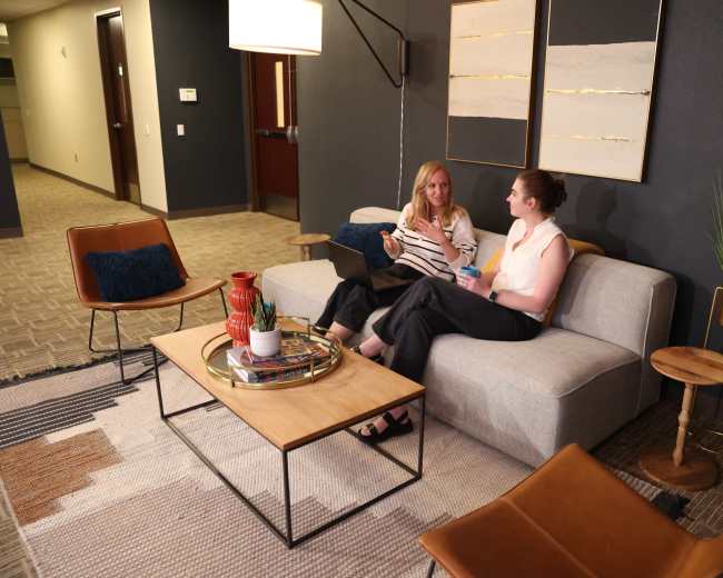 Two women sit on a light gray sofa in a modern office lounge area, engaged in conversation while a laptop rests on the coffee table between them.