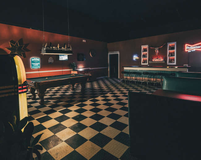 The image depicts a dimly lit game room featuring a pool table, a retro jukebox, and a bar counter with various neon signs on the walls.