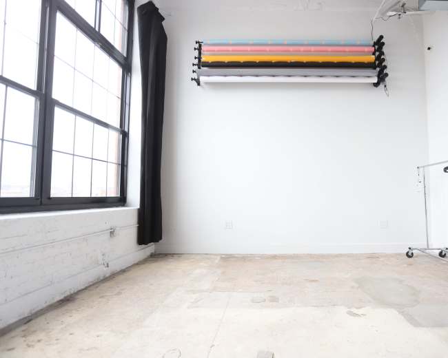 The image shows a spacious, empty room with large windows and a clothing rack on wheels against a white wall.