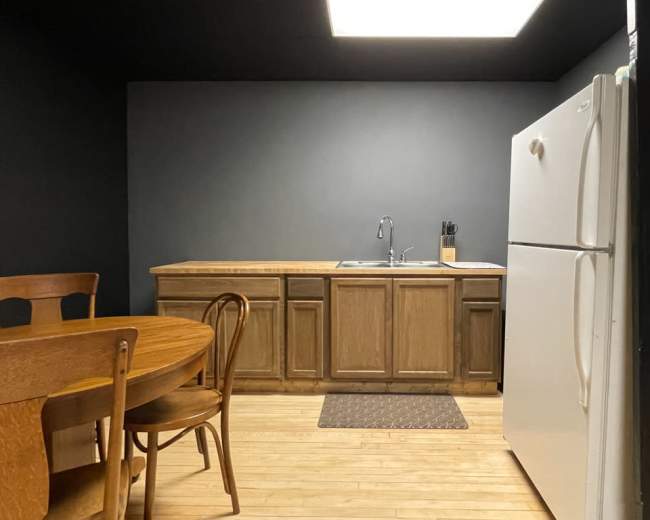 A simple kitchen features a wooden table with chairs, a countertop with a sink, and a white refrigerator against a black wall.