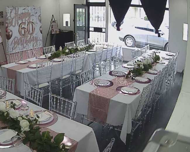 The image shows a decorated event space with two long tables set for a celebration, featuring white tablecloths, pink accents, and floral arrangements along the center of each table.