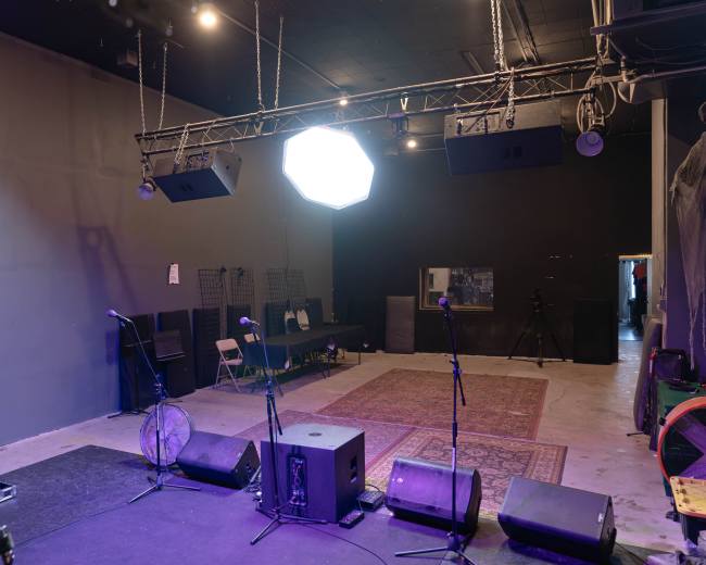 The image shows a performance space with a stage featuring two microphones, speakers, and a rug on the floor, along with chairs and equipment in the background.