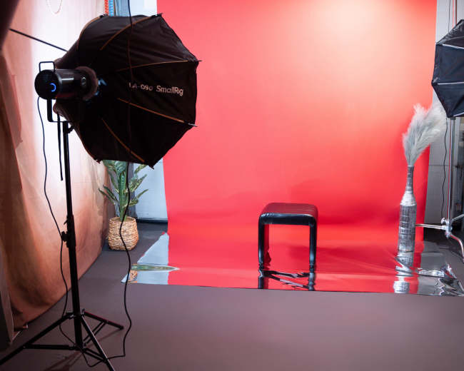 The image displays a photography studio setup with two softbox lights, a red backdrop, a black stool in the center, and a decorative plant in a woven basket to the left.