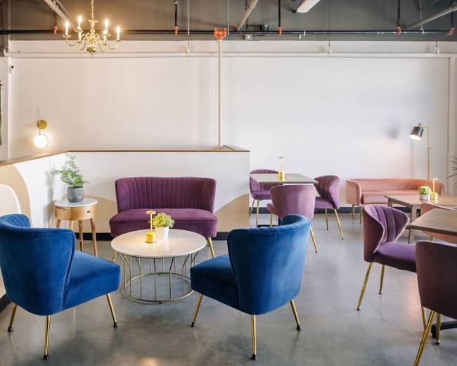 The image shows a modern lounge area with purple and blue upholstered chairs, a round table, and a small potted plant, set against a neutral wall and ceiling with industrial lighting.