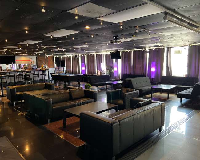 The image shows a spacious lounge area with black leather furniture, tables, and soft lighting, featuring a bar at one end.