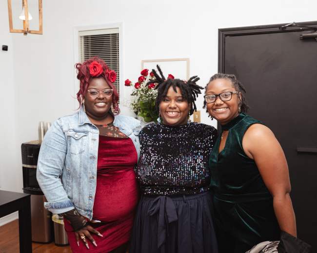 Three women pose together indoors, each wearing stylish outfits, with a backdrop of flowers and decorative elements.