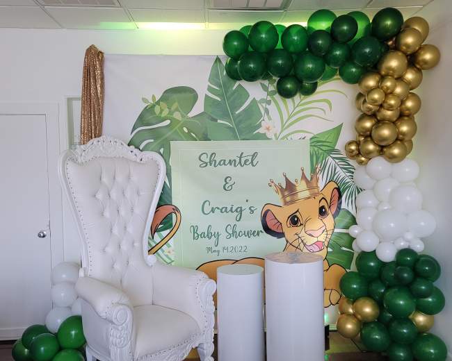 The image shows a decorative backdrop for a baby shower featuring a white throne chair, green and gold balloons, and a sign with the names "Shantel & Craig" along with a Lion King theme.
