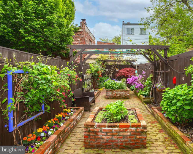 The image shows a landscaped garden pathway bordered by raised brick planters filled with vibrant flowers and greenery, set against wooden fencing.