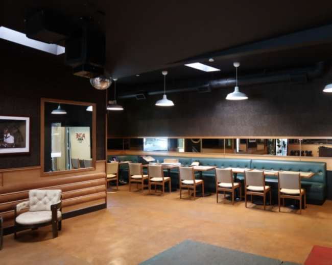 The image shows a modern interior of a restaurant or café with a seating area featuring a long green bench and several tables with chairs, illuminated by pendant lights.