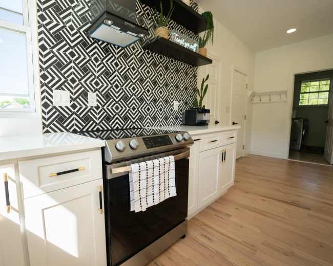 A modern kitchen features a black and white geometric patterned backsplash, a range with a towel hanging from its oven handle, and stylish cabinets with gold hardware.