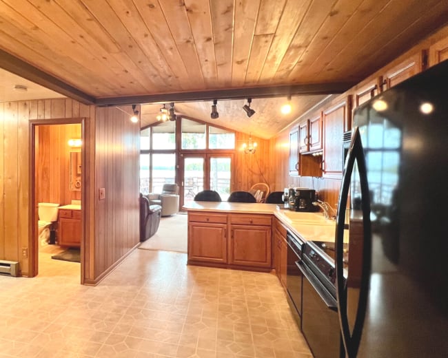 A wooden-finished kitchen with a view into a sunlit living area, featuring a refrigerator, stove, and cabinetry.