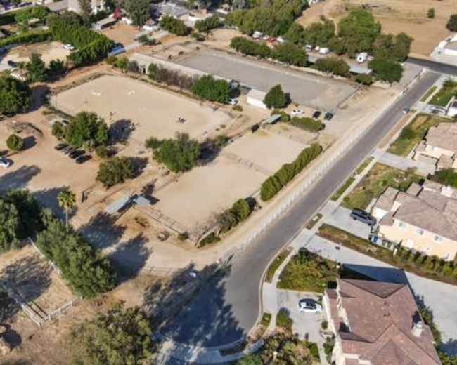 The image shows an aerial view of a rural property with an equestrian arena, surrounded by houses and trees, along with nearby roads.
