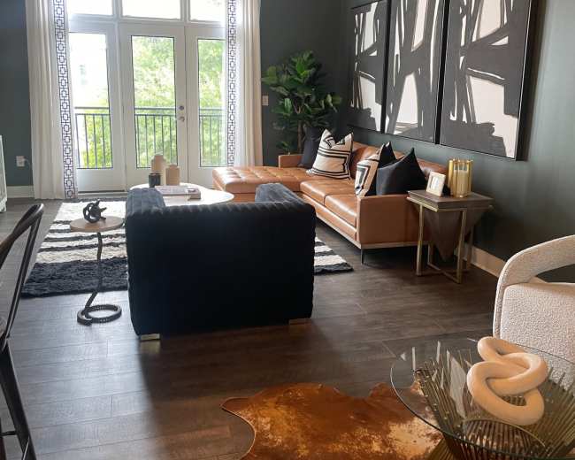 The image shows a modern living room with a leather sofa, a black velvet chair, and a decorative glass coffee table on a cowhide rug, along with large windows that allow natural light to enter the space.