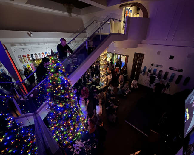 A crowded indoor event features a large Christmas tree adorned with colorful lights, with attendees gathered around a presentation on a screen.