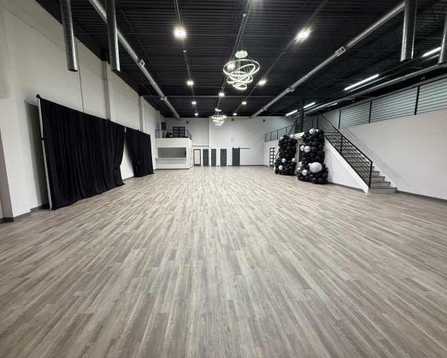 The image shows a spacious, empty room with wooden flooring, high ceilings, and minimalist decor, featuring a staircase on one side and black curtains on another.