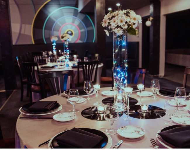 A round dining table is set with black napkins and glassware, adorned with a tall floral centerpiece and illuminated by soft lighting in a modern restaurant.