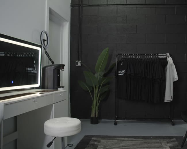 The image shows a minimalist interior space with a clothing rack, a plant, a chair, and a mirror against a black wall.
