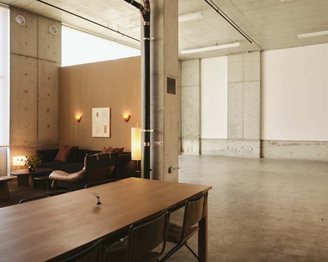 The image shows a spacious, industrial-style room with concrete walls, a seating area on one side, and an empty section suitable for events or presentations.