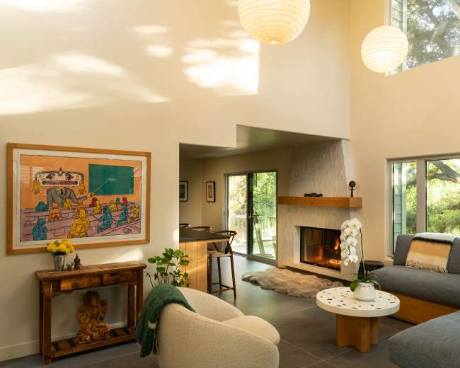 A modern living room features a fireplace, large windows, and a mix of artwork and furniture, creating an open and inviting space.