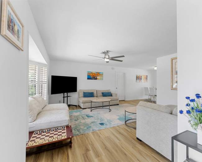 The living room features light-colored sofas, a coffee table, and a large area rug, with a television in the corner and a vase of flowers on a side table.
