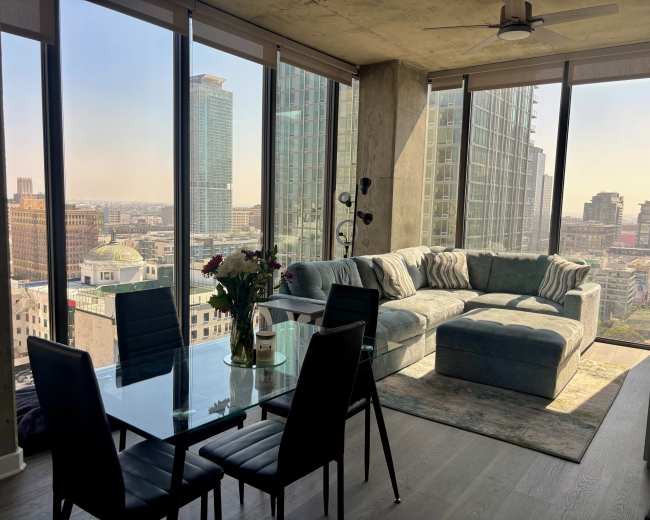A modern living area features a glass dining table and a sectional sofa, with large windows providing a view of the city skyline.