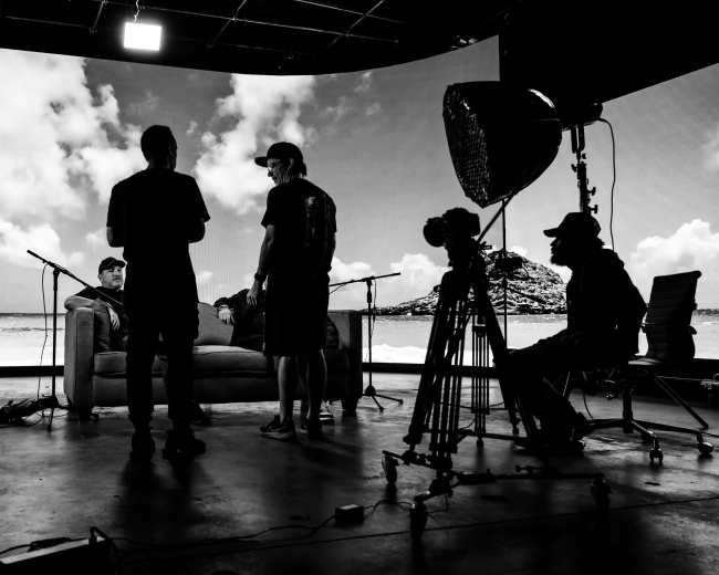 A film production crew gathers around a sofa on a set with a large backdrop displaying clouds and a beach scene.
