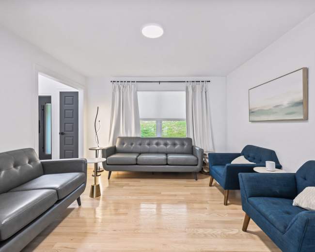 A modern living room features a gray leather couch, two blue armchairs, a floor lamp, and large windows with light curtains.