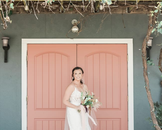 A bride stands in front of a pink double door, holding a bouquet, with her lace-trimmed gown trailing on the ground.